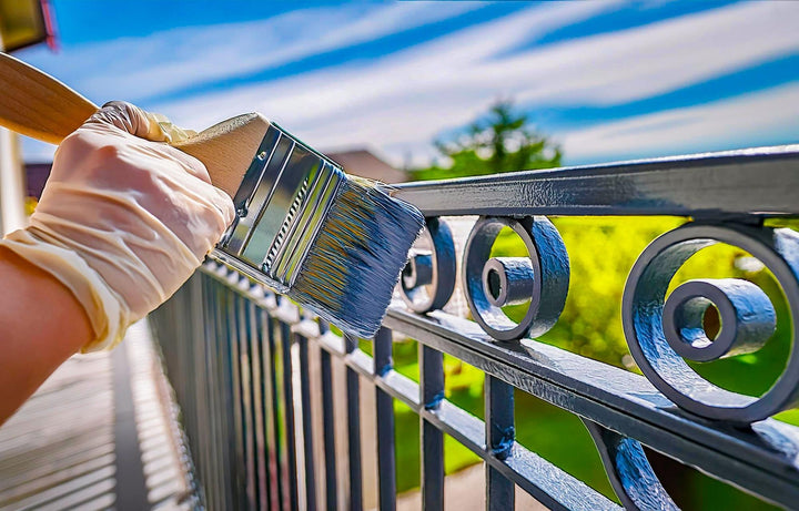 Hands applying paint with a brush to a metal porch railing above a drop cloth